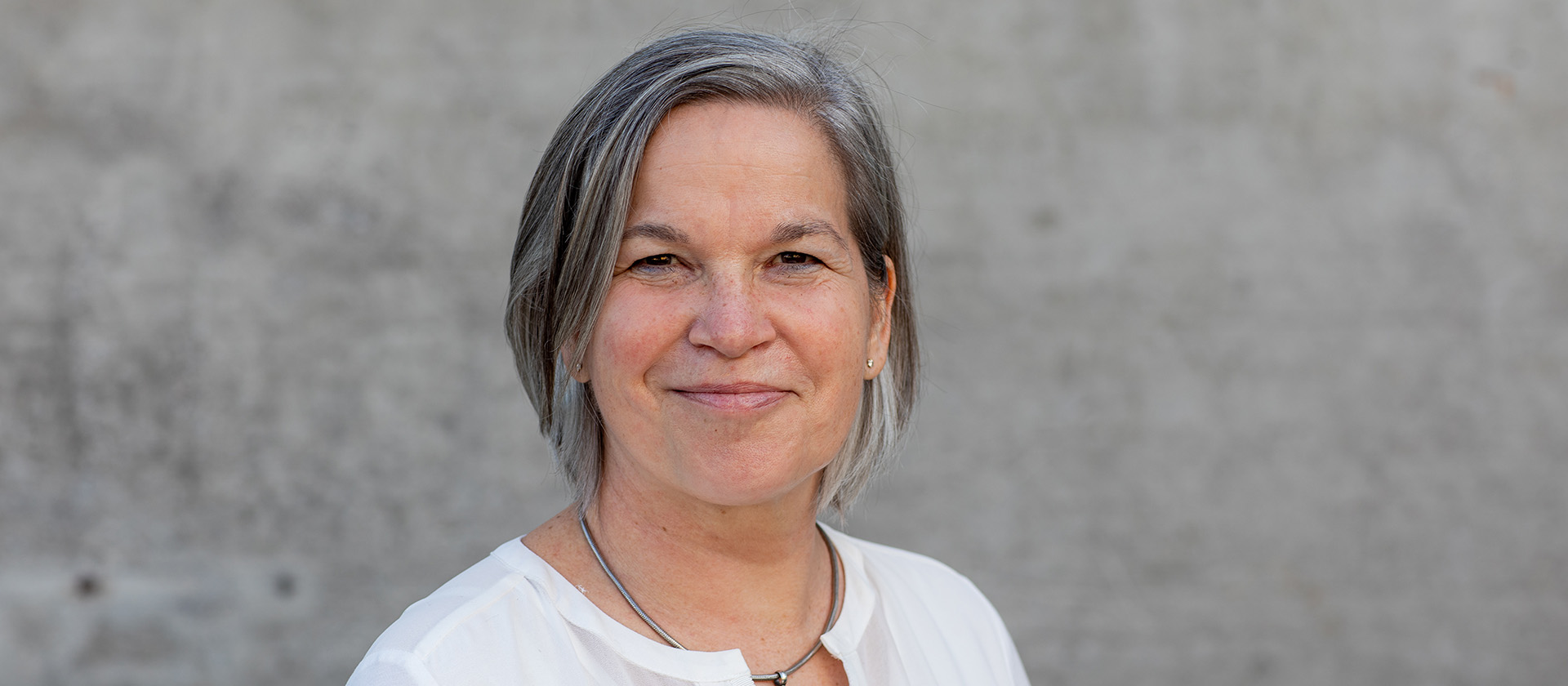 Porträtfoto Antonia Engel mit grauen kurzen Haaren, dunklen Augen und weißem Oberteil. Sie steht vor einer grauen Wand und sieht freundlich den Betrachter an.