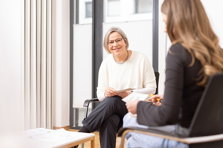 Frau mit grauen, kurzen Haaren in schwarzer Hose und weißem Oberteil sitzt auf einem Stuhl mit Armlehnen. Sie schaut zu einer Person die rechts im Bild ebenfalls auf einem Stuhl sitzt und auf einen Zettel in ihrer linken Hand schaut.