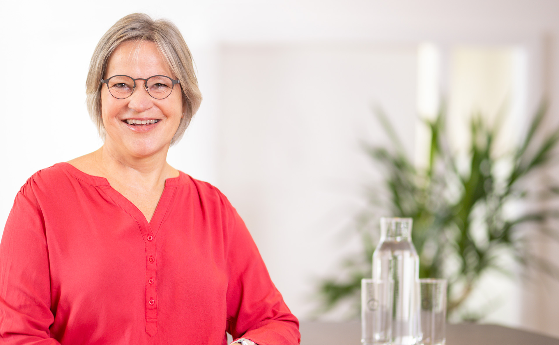 Frau mit grauen kurzen Haaren, schwarzer Brille und rotem Oberteil schaut den Betrachter an. Sie stützt sich mit ihrem linken Arm auf einen Tisch links neben ihr, auf dem zwei Gläser und eine Wasserkaraffe aus Glas stehen.